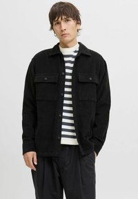 Jeune homme portant une veste en velours côtelé noire sur une chemise à rayures blanches et noires, se tenant avec une main dans sa poche contre un fond uni.