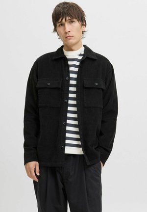Young man wearing a black corduroy jacket over a white and black striped shirt, standing with one hand in his pocket against a plain background.