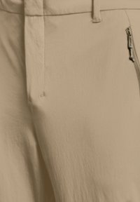 Primo piano di pantaloni beige con tasca laterale con cerniera e dettaglio passante per cintura su tessuto testurizzato.