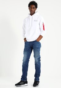 Witte hoodie met een logo aan de voorkant, rode rits aan de zijkant, blauwe slim-fit jeans en zwarte sportschoenen met witte accenten.
