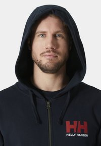 Sudadera con cremallera azul marino hecha de algodón, con una gran capucha, cremallera frontal y un logo rojo en el lado izquierdo del pecho. Textura suave.