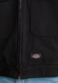 Sort, kraftig stofjakke med frontlynlås og to klaplommer. Har et syet "Dickies" logo nederst til venstre.