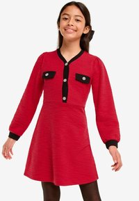 Rotes Kleid mit strukturierter Oberfläche, schwarzem Akzent und zwei Brusttaschen. Hat lange Ärmel und ist knielang mit Knopfdetails.