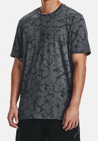 Grå sportt-shirt med ett svart abstrakt mönster, korta ärmar, rund hals och Under Armour-logotyp. Tillverkad av ett slätt, lätt material.