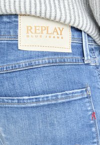 Svijetloplave traperice sa kožnom etiketom s natpisom "REPLAY BLUE JEANS," sa detaljima šavova i vidljivim stražnjim džepom.