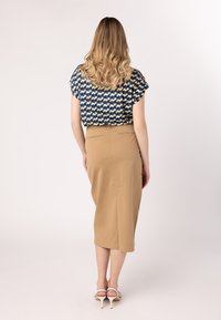 Transparante blauwe blouse met een geel en wit patroon, gecombineerd met een beige midi-rok. De rok heeft een split aan de achterkant en minimale hardware.