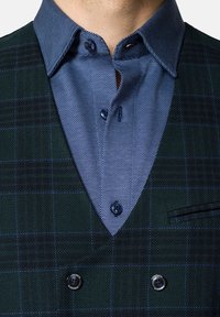 Zielona kamizelka w kratę z niebieską wzorzystą koszulą. Cechy to dekolt w serek, niebieskie guziki oraz mała kieszeń po lewej stronie.