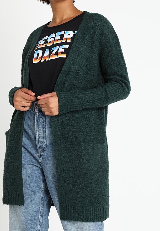 Femme portant un cardigan vert foncé ouvert sur un t-shirt noir "Desert Daze" et un jean taille haute bleu clair, les mains tenant les bords du cardigan.