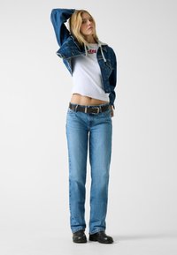 Veste en denim avec un T-shirt blanc court, jean droit bleu clair, ceinture noire et chaussures noires. La veste a une coupe classique et des boutons.