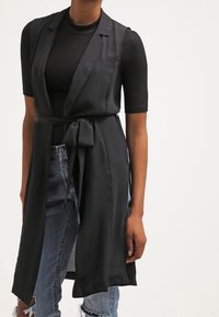 Frau in einem schwarzen ärmellosen, taillierten Weste über einem schwarzen gerippten T-Shirt mit kurzen Ärmeln und verwaschenen blauen Jeans vor einem einfarbigen Hintergrund.