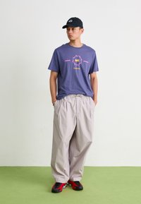 Violet grafiko marškiniai su rožiniu ir geltonu Nike logotipu, derinami su šviesiai pilkomis platėjančiomis kelnėmis ir juodais/raudonais sportbačiais. Paprasta juoda kepurė.