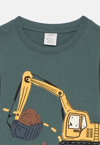 Τιρκουάζ βαμβακερό T-shirt με κίτρινο γραφικό εκσκαφέα που σηκώνει έναν καφέ σωρό χώματος, σχεδιασμένο με μαύρα περιγράμματα και παιχνιδιάρικες λεπτομέρειες.