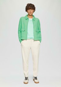 Veste texturée verte sur une chemise à rayures blanches et vertes, associée à un pantalon de survêtement crème et des baskets blanches avec des accents noirs.