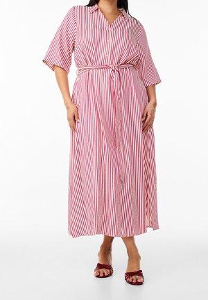 Femme portant une robe midi rayée verticale rouge et blanche, boutonnée, avec une ceinture nouée à la taille et des manches courtes, associée à des sandales rouges à bout ouvert.