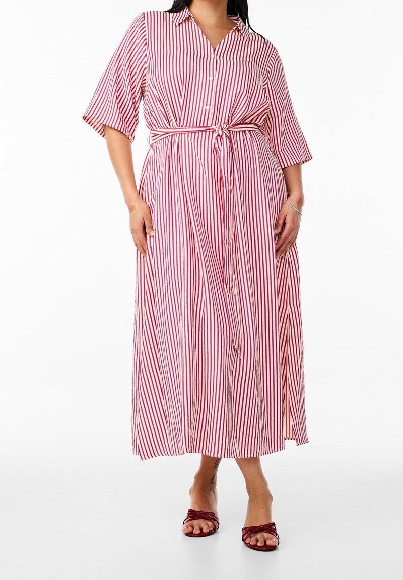 Femme portant une robe midi rayée verticale rouge et blanche, boutonnée, avec une ceinture nouée à la taille et des manches courtes, associée à des sandales rouges à bout ouvert.