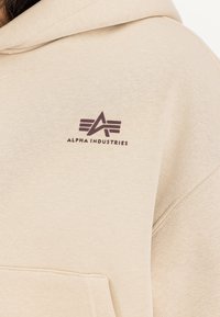 Bēša kapuce, izgatavota no mīksta materiāla, ar priekšu kabatu un tumši brūnu logotipu kreisajā krūtī ar stilizētu fontu.