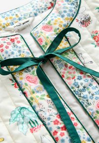 Gequilte kleding met meerkleurige bloemmotieven, donkergroen detail, gestrikte stoffen strik en kleine stripfiguren op een witte achtergrond.