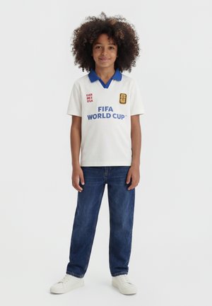 Barn med krøllet hår iført en hvid FIFA World Cup polo, blå jeans og hvide sneakers, stående mod en ensfarvet baggrund.