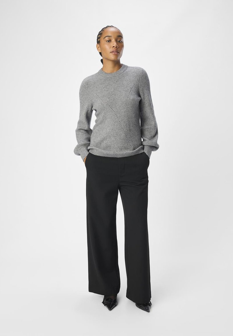 Szary sweter z dzianiny o ribbowej strukturze i diagonalnym wzorze, zestawiony z czarnymi szerokimi spodniami i czarnymi, spiczastymi butami.