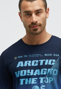 Navyblaue T-Shirt mit auffälliger blauer Typografie und dem Text "ARCTIC VOYAGE TO THE TOP" sowie einem grafischen Segeldesign.