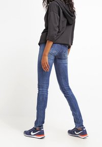 Wąskie dżinsy w średnim niebieskim kolorze z detalami w postaci przetarć, połączone z czarną bluzą z kapturem oraz granatowymi sneakersami z pomarańczowymi akcentami.