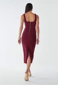 Burgunderfarvet tætsiddende kjole med firkantet ryg, lynlås og slids foran. Glat tekstur og struktureret design. Sammen med strappy guld hæle.