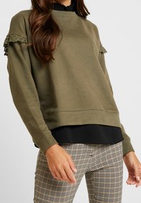 Mujer que lleva un suéter oliva con encaje en las mangas, camiseta negra debajo y pantalones de cuadros beige, de pie contra un fondo plano.