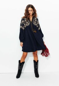 Robe bleu marine avec des motifs floraux brodés, manches bouffantes longues, décolleté en V et détails à franges. Portée avec des bottes de cow-boy noires et un sac à franges.