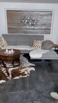 Sofá seccional cinza com almofadas estampadas, mesa de café circular em metal preto e marrom, e um tapete de pele de vaca sobre o piso de azulejos escuros.