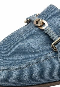 Mocassin en tissu texturé bleu avec détail en chaîne dorée sur le dessus et couture visible sur le côté.