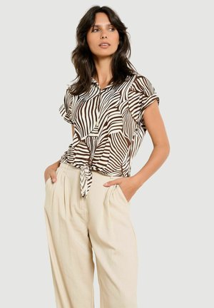 Femme aux longs cheveux foncés portant une chemise à manches courtes à rayures zébrées marron et blanches boutonnée, nouée à la taille, et un pantalon beige taille haute.