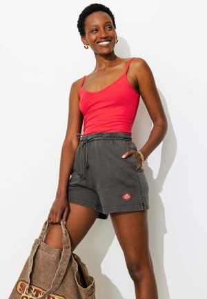 Femme souriante en débardeur rouge et short gris tenant un grand sac cabas marron, debout sur un fond blanc.