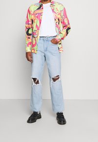Camisa de botones colorida con patrones abstractos en rosa, verde y amarillo, combinada con jeans rotos de un azul claro y zapatos negros.