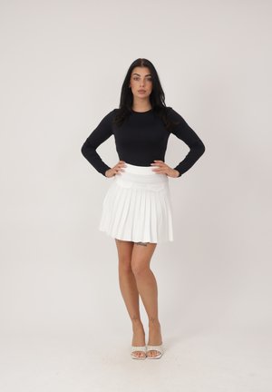 Frau mit langen dunklen Haaren, die ein schwarzes Langarmshirt, einen weißen plissierten Rock und weiße offene High Heels trägt, stehend mit den Händen in den Hüften.