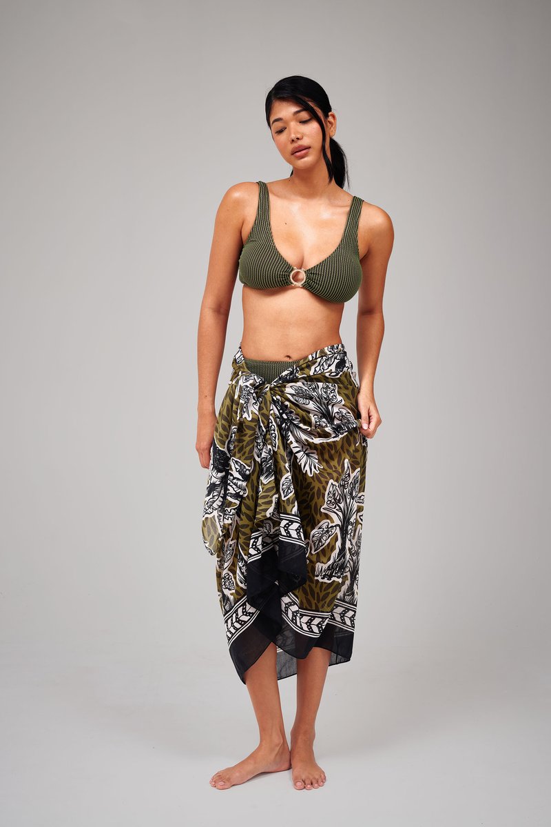 Vrouw staat op blote voeten en draagt een olijfgroene bikini top met ringdetail en een patroonrijke wikkelrok tegen een effen achtergrond.