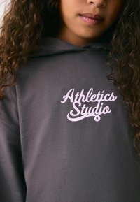 Grå hættetrøje med en blød tekstur, der har et hvidt skrifttype-logo "Athletics Studio" tydeligt vist på forsiden.