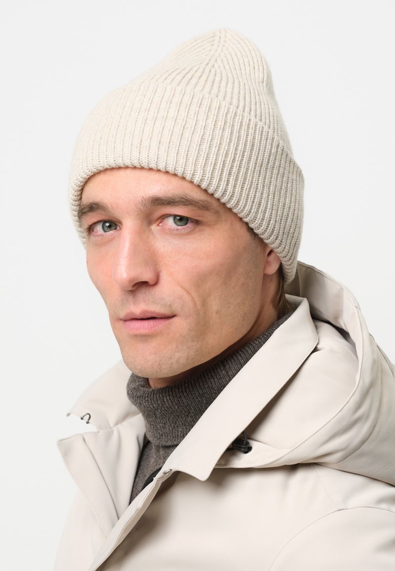 Gorro de punto beige con canalé vertical, de punta, llevado con un suéter gris de cuello alto y chaqueta de color claro. Fondo liso.
