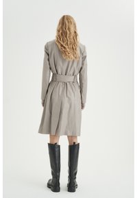 Jasnoszary płaszcz trench z paskiem w talii, tylnym wentylatorem i długimi rękawami, w połączeniu z czarnymi skórzanymi botkami do kolan. Gładka tekstura, minimalistyczny design.