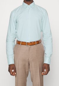 Camicia button-up azzurro chiaro con colletto, bottoni bianchi e logo ricamato, abbinata a pantaloni beige e una cintura marrone.