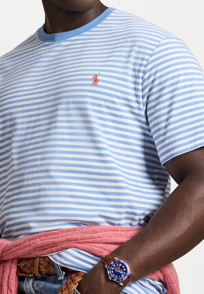 Svītrainā T-kreklā gaiši zilā un baltā krāsā, ar apaļu kakla izgriezumu un sarkanu logo. Valkāta kopā ar rozā apmetni un brūnu jostu, demonstrējot zilu pulksteni.