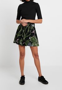 Haut noir à manches courtes associé à une jupe noire évasée avec un motif botanique vert et rose ; le mannequin porte des baskets noires.