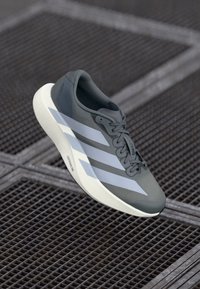 Zapatilla deportiva gris con tres franjas diagonales blancas, un diseño elegante, textura suave y una suela blanca acolchada.