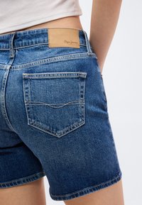 Οπίσθια όψη προσώπου που φοράει μπλε τζιν σορτς με ραμμένη πίσω τσέπη και καφέ δερμάτινη ετικέτα Pepe Jeans στη μέση.