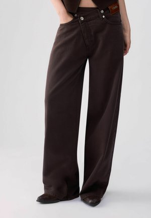 Personne portant un jean marron foncé taille haute à jambes larges avec des chaussures noires pointues, une main dans une poche, sur un fond uni.