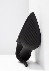 Escarpin en suede noir à bout pointu et talon aigu mince. La semelle présente un motif texturé pour l'adhérence. Détail de zip sur le côté.