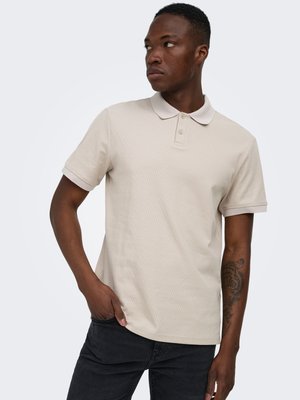 Ung mand med tatovering på venstre underarm iført en beige kortærmet polo og sorte jeans, der kigger til siden med den ene hånd i lommen.