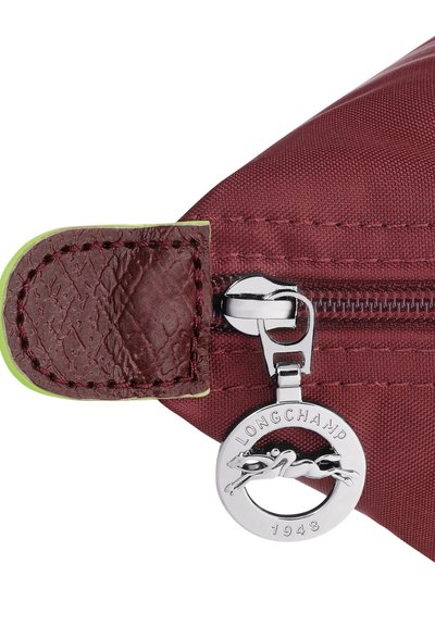 Bordeauxfarbene Reissverschluss-Tasche mit strukturierten Lederelementen und einem silbernen, runden Logo, das ein springendes Pferd zeigt. Grüner Reißverschluss-Zug sichtbar.