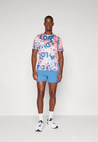 Krāsains, īsām piedurknēm T-krekls ar rozā un zilā abstraktā rakstā un tekstu; apvienots ar zilām šortiem un baltām sporta apaviem.