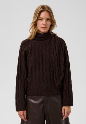 Femme blonde portant un pull texturé marron foncé avec un détail d'écharpe et un pantalon en cuir marron foncé, debout devant un fond uni.