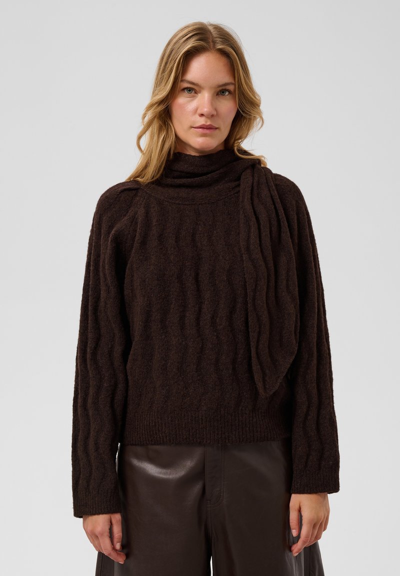 Femme blonde portant un pull texturé marron foncé avec un détail d'écharpe et un pantalon en cuir marron foncé, debout devant un fond uni.